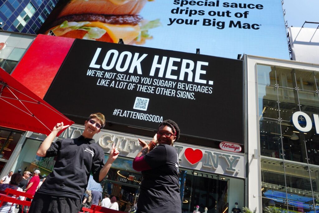 TFFJ alums Fiona C. and Najae S. posing in front of the #FlattenBigSoda ad in Times Square.